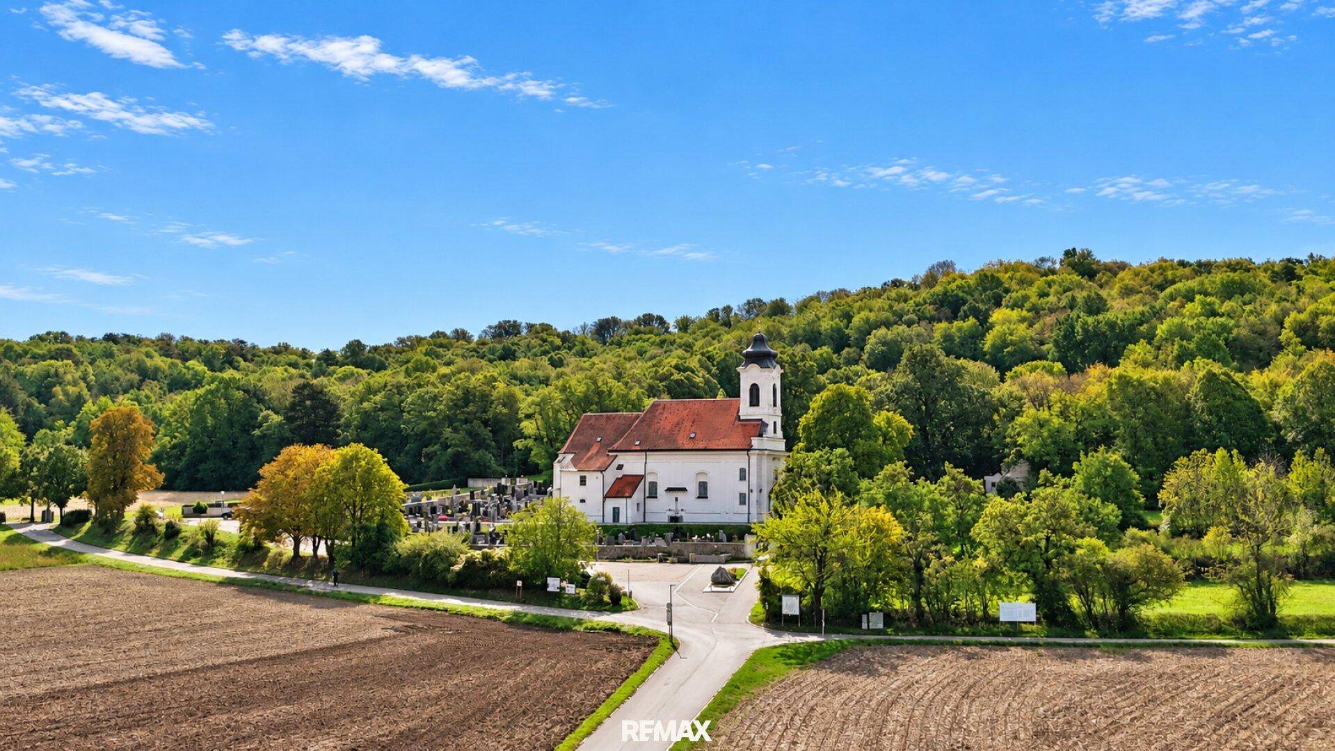 Luftaufnahme einer historischen Kirche mit Friedhof, umgeben von üppiger Natur und Feldern.