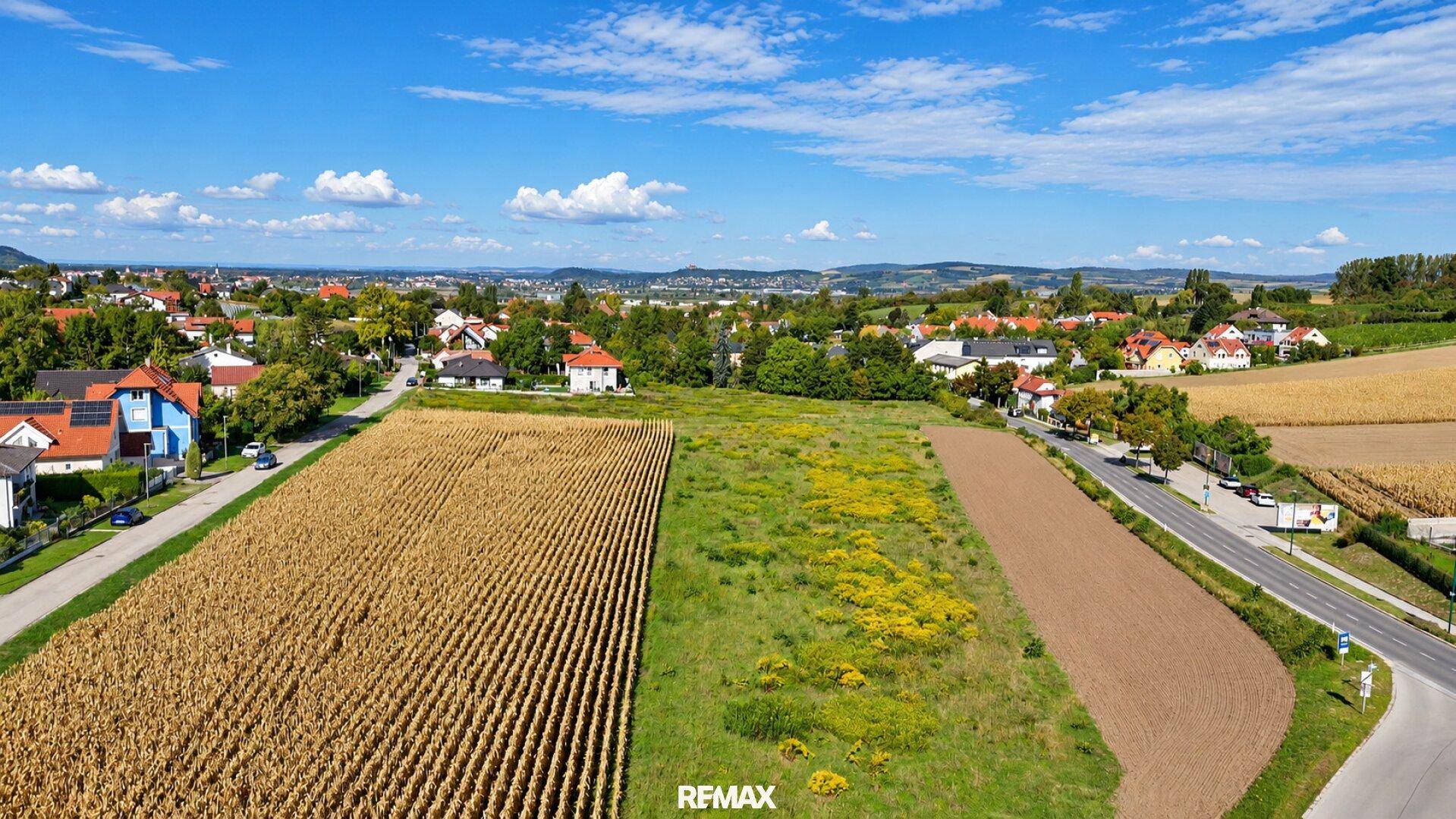 Weitläufige Luftaufnahme einer Siedlung mit Feldern und Wohnhäusern in ländlicher Umgebung.