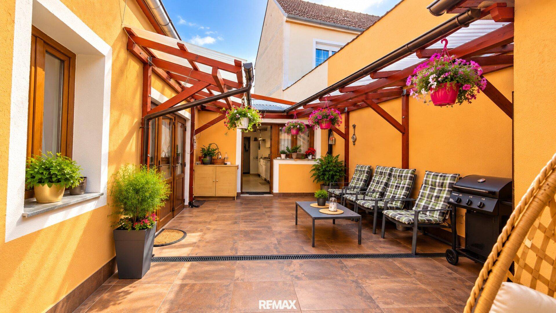 Sonnige Terrasse mit Grillbereich, bequemen Sitzgelegenheiten und farbenfrohen Blumenampeln.