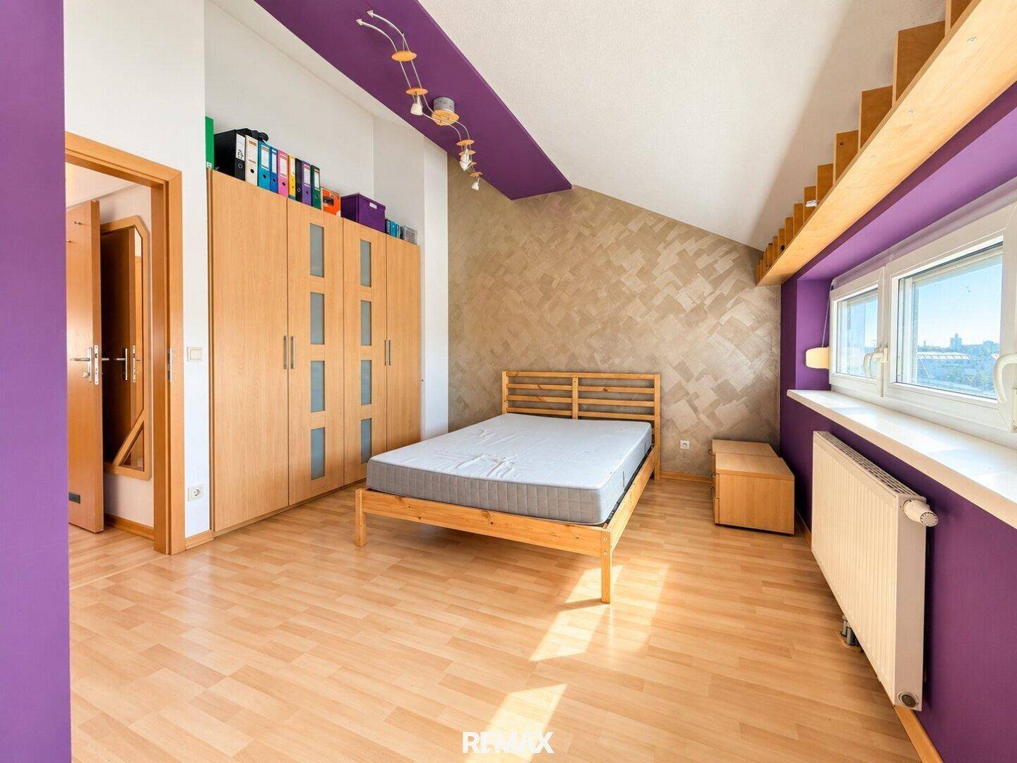 Schlafzimmer mit lila Wänden, Holzbett, Einbauschrank und schräger Decke.