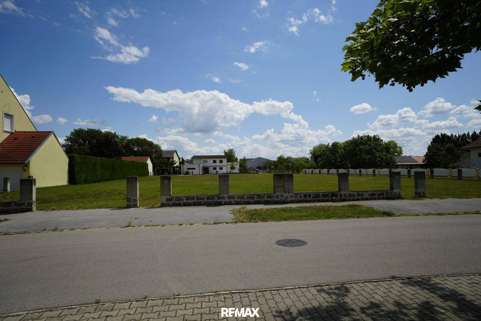 Weitläufiges Grundstück mit grüner Wiese, umgeben von einer Straße und Wohnhäusern unter blauem Himmel.