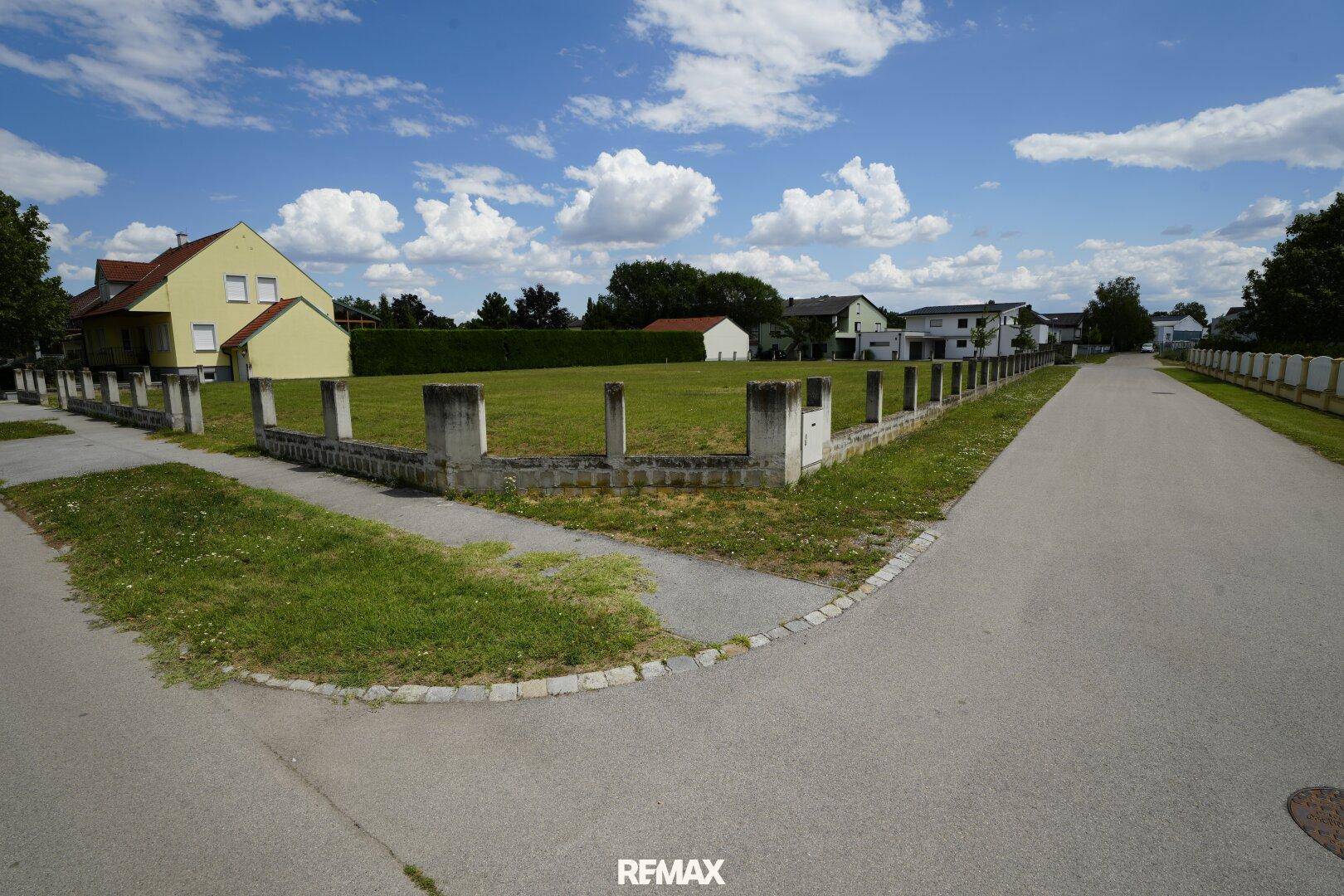 Blick auf ein unbebautes Grundstück an einer Straßenecke mit niedriger Betonmauer und umliegenden Häusern.