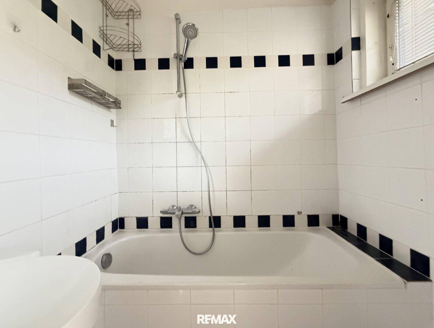 Badezimmer mit Badewanne, Duschvorrichtung und weißen Fliesen mit schwarzem Akzentstreifen.