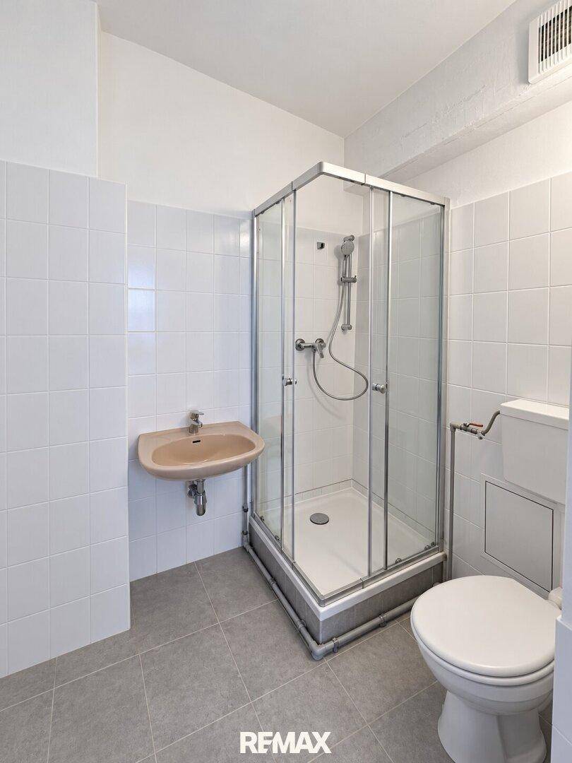 Badezimmer mit Eckdusche, Toilette, kleinem Waschbecken und weißen Wandfliesen.