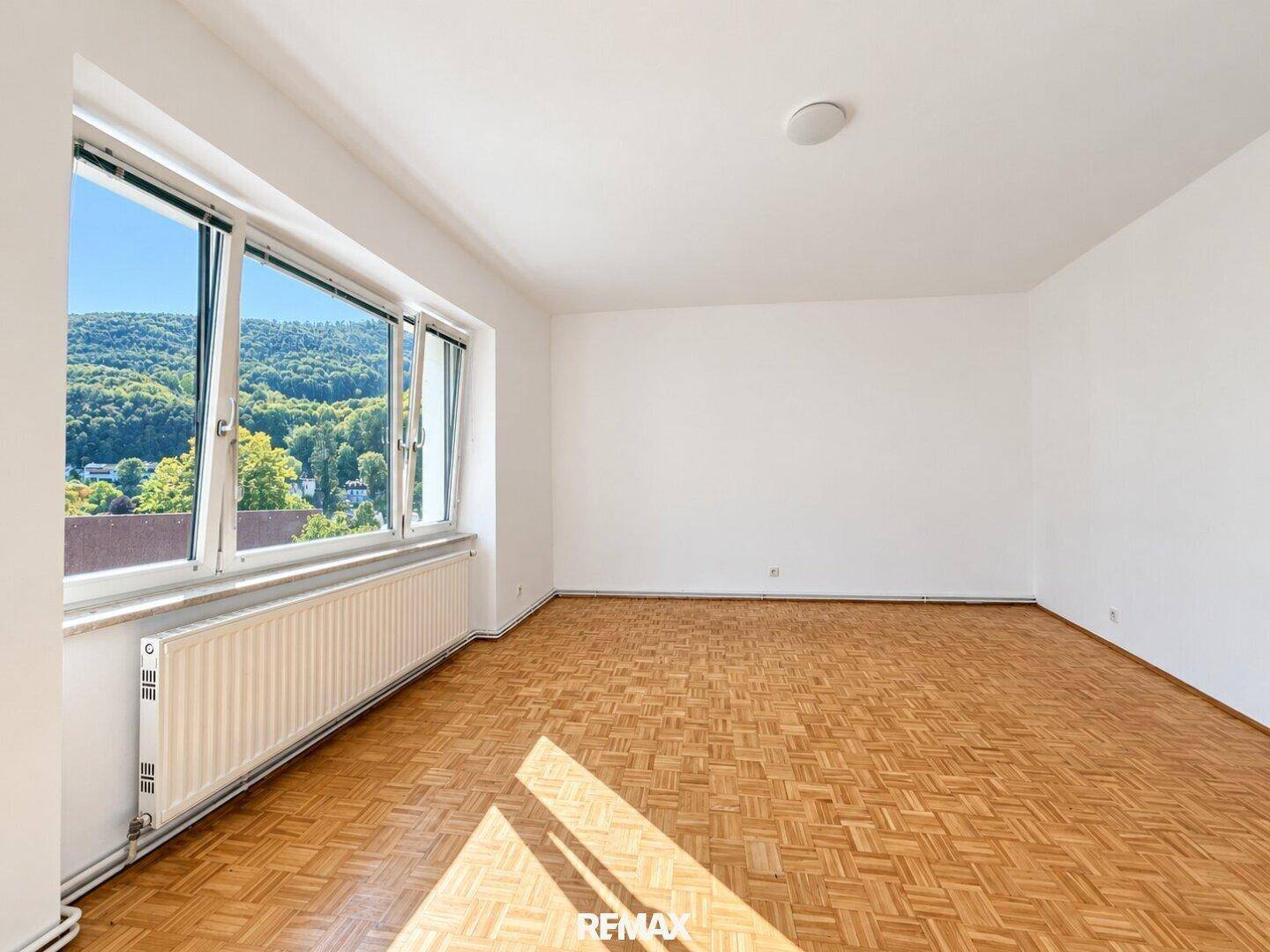 Heller Raum mit Parkettboden, weißen Wänden und großem Fenster mit grüner Aussicht.