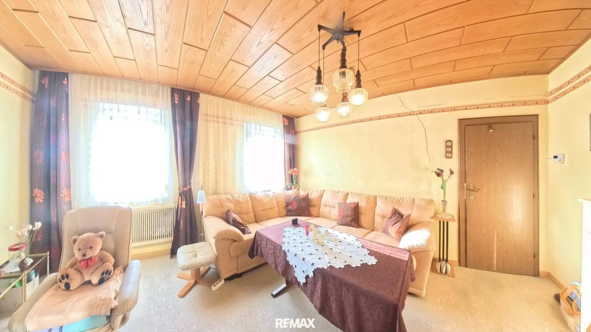 Wohnzimmer mit beiger Eckcouch, Holztäfelung an der Decke und Teppichboden.