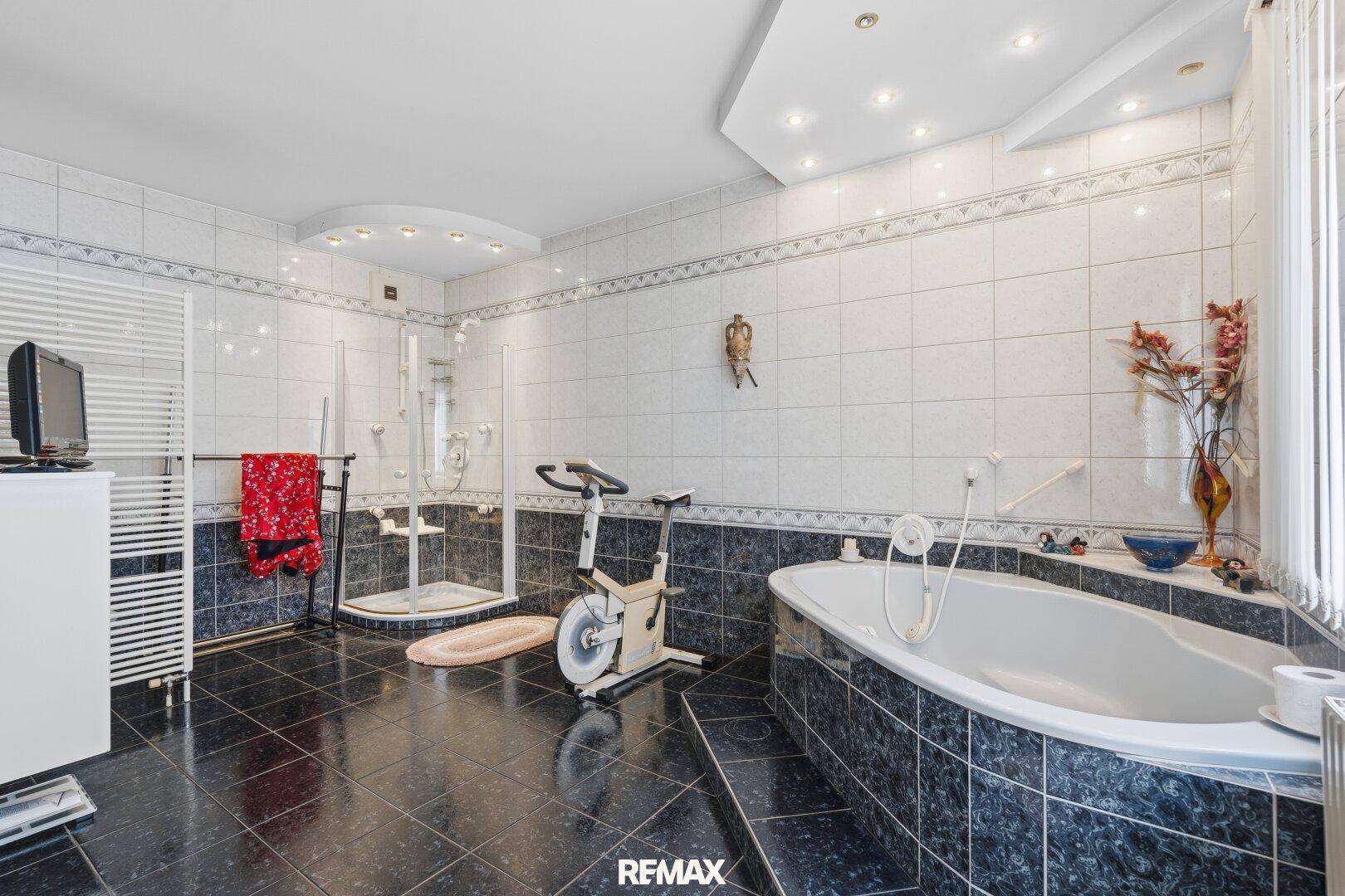 Großes Badezimmer mit Badewanne, Dusche, Fitnessgerät und dunklen Fliesen.