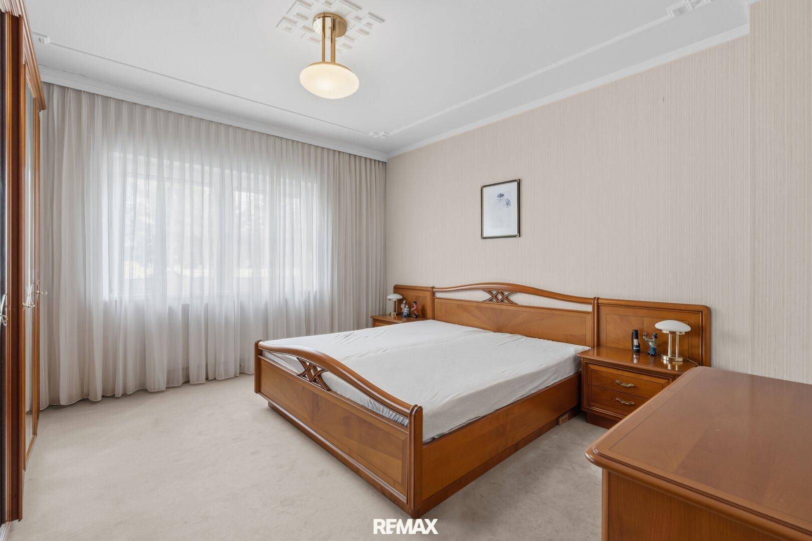 Schlafzimmer mit Doppelbett, Teppichboden und großem Fenster mit Vorhängen.