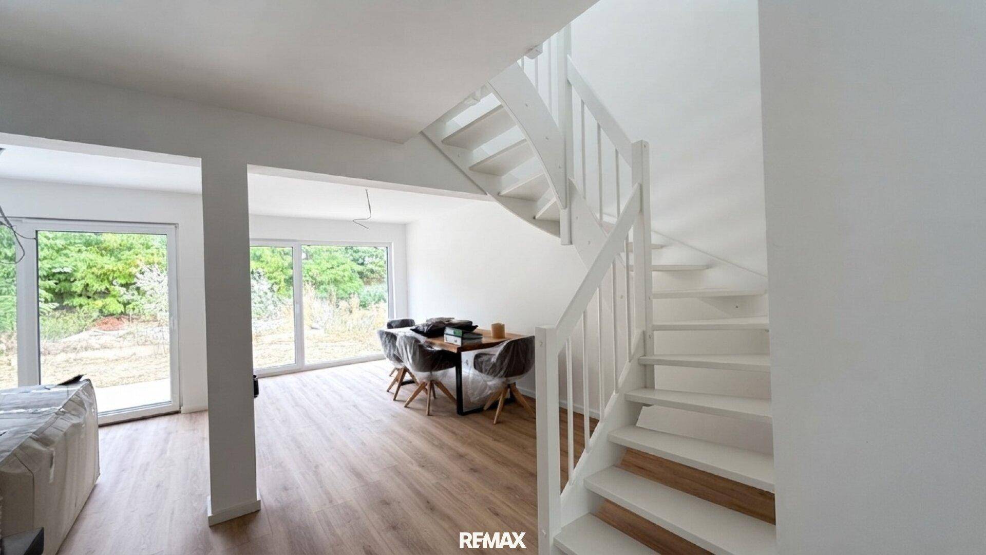 Heller Wohn- und Essbereich mit Laminatboden, großen Fenstern und einer modernen Holztreppe.