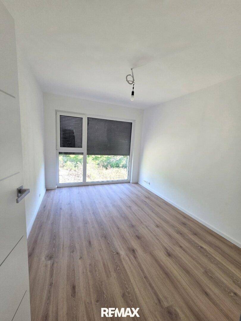 Leeres Zimmer mit Laminatboden, weißen Wänden und großem Fenster mit Rollladen.