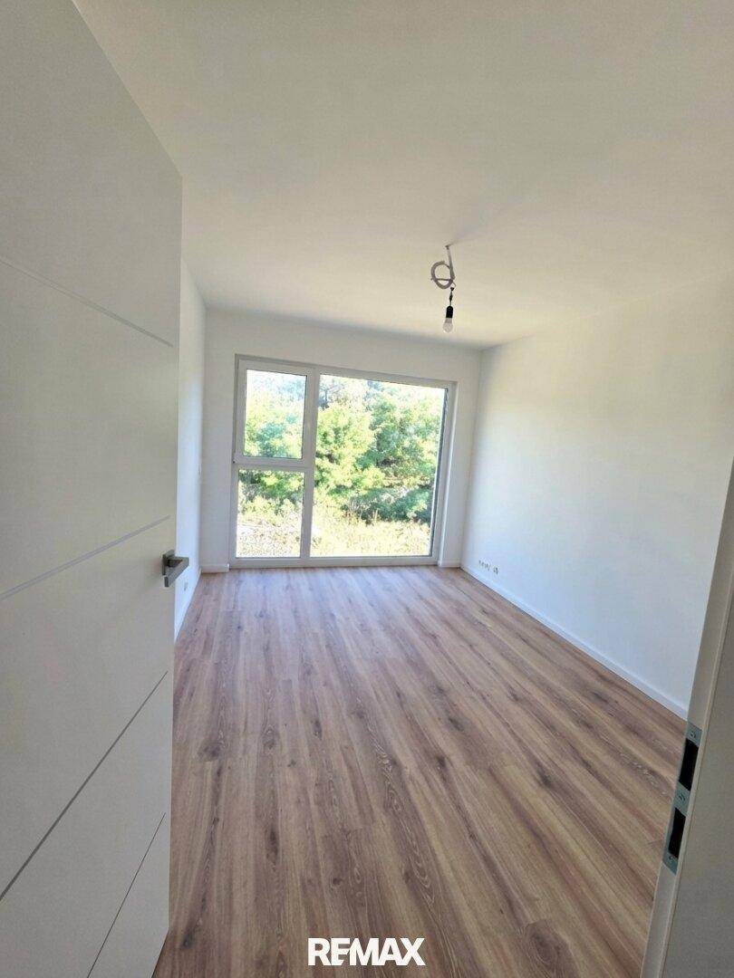 Helles Zimmer mit Laminatboden, weißen Wänden und Blick ins Grüne durch das große Fenster.
