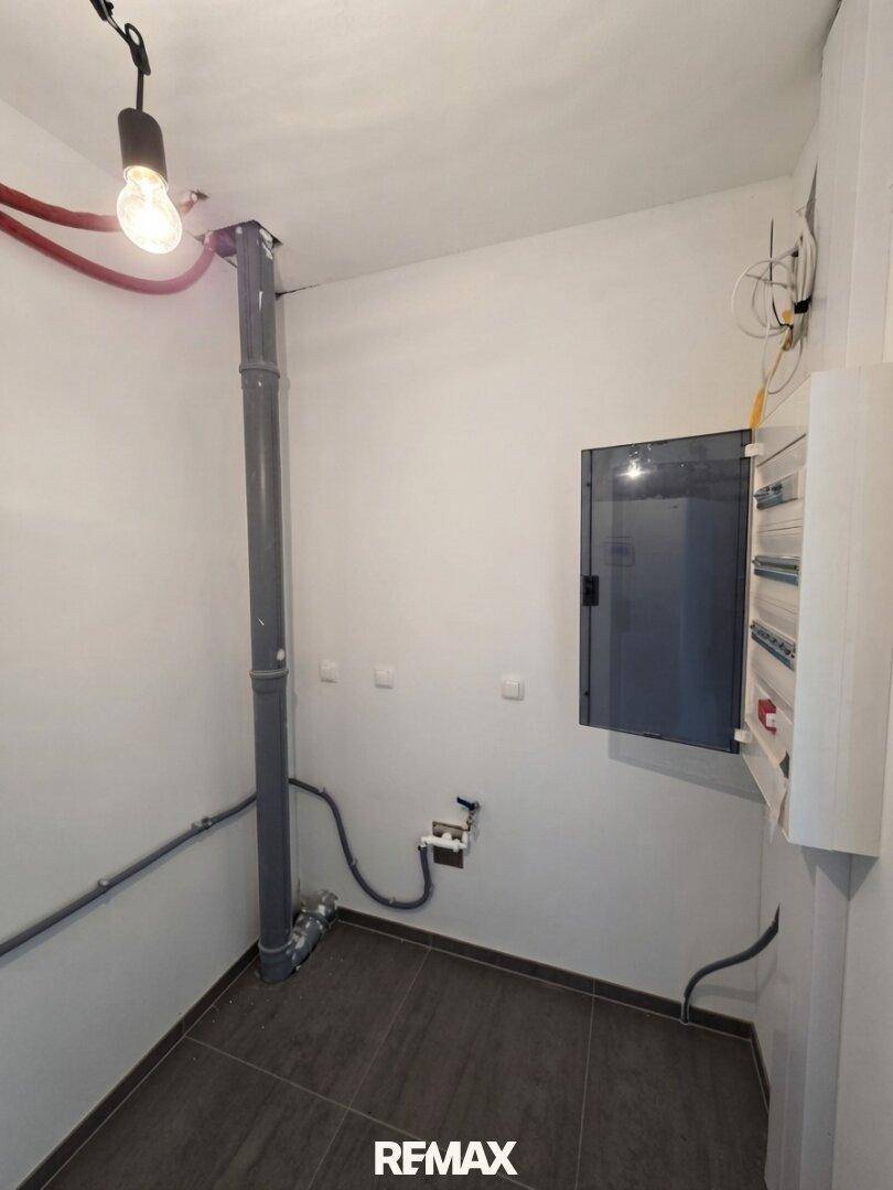 Technikraum mit sichtbaren Installationen und einem Sicherungskasten auf gefliestem Boden.