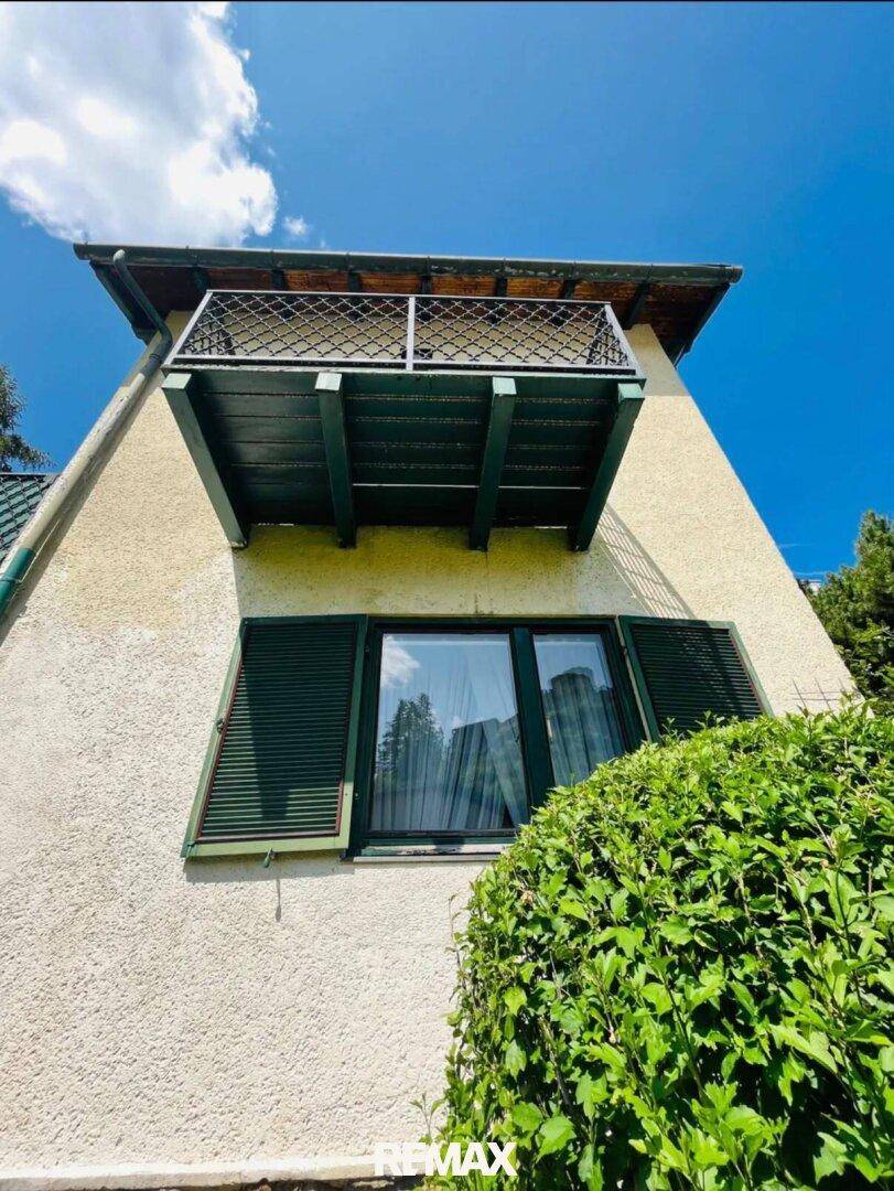 Balkon an der Hausfassade mit schmiedeeisernem Geländer und grünen Fensterläden unter blauem Himmel.