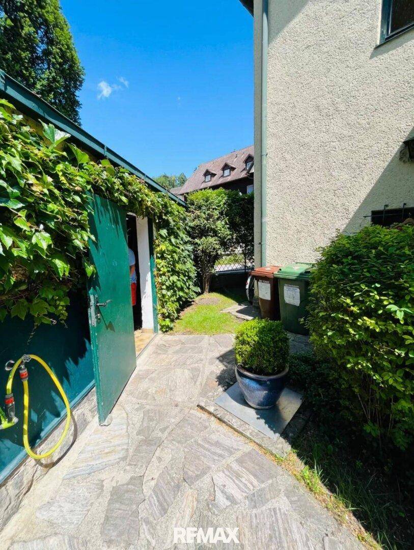 Gepflasterter Weg entlang des Hauses mit üppigem Grün und einer grünen Tür, die zu einem Nebengebäude führt.