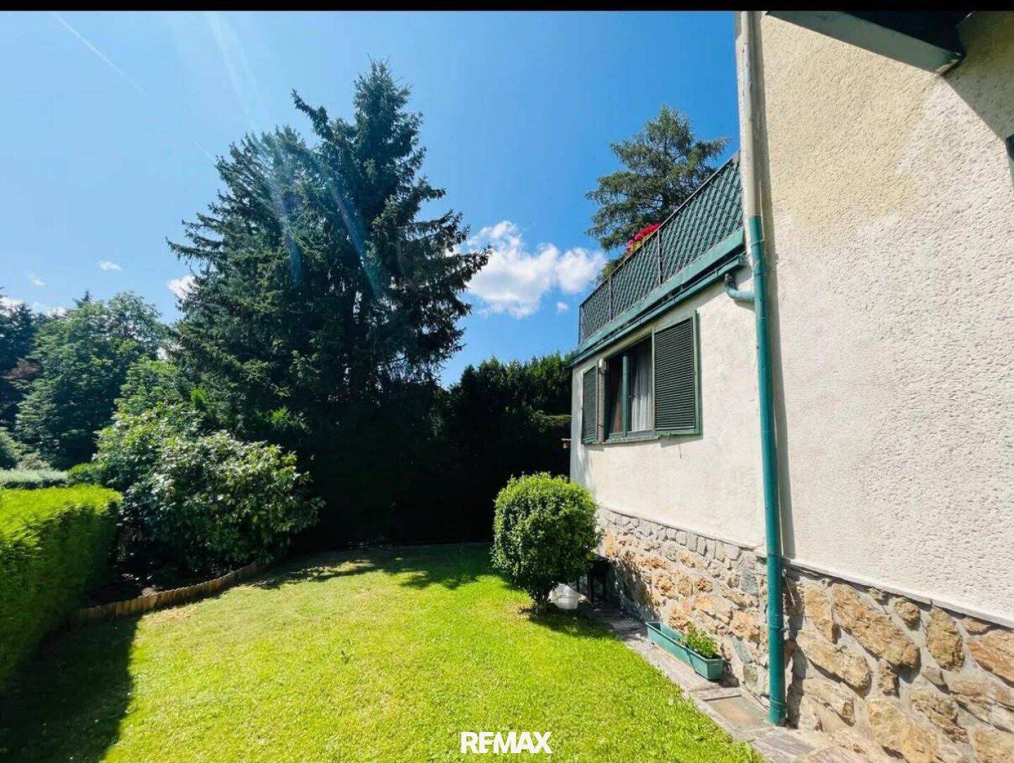 Seitenansicht des Hauses mit gepflegtem Rasen, hohen Bäumen und einer Steinmauer im unteren Bereich.
