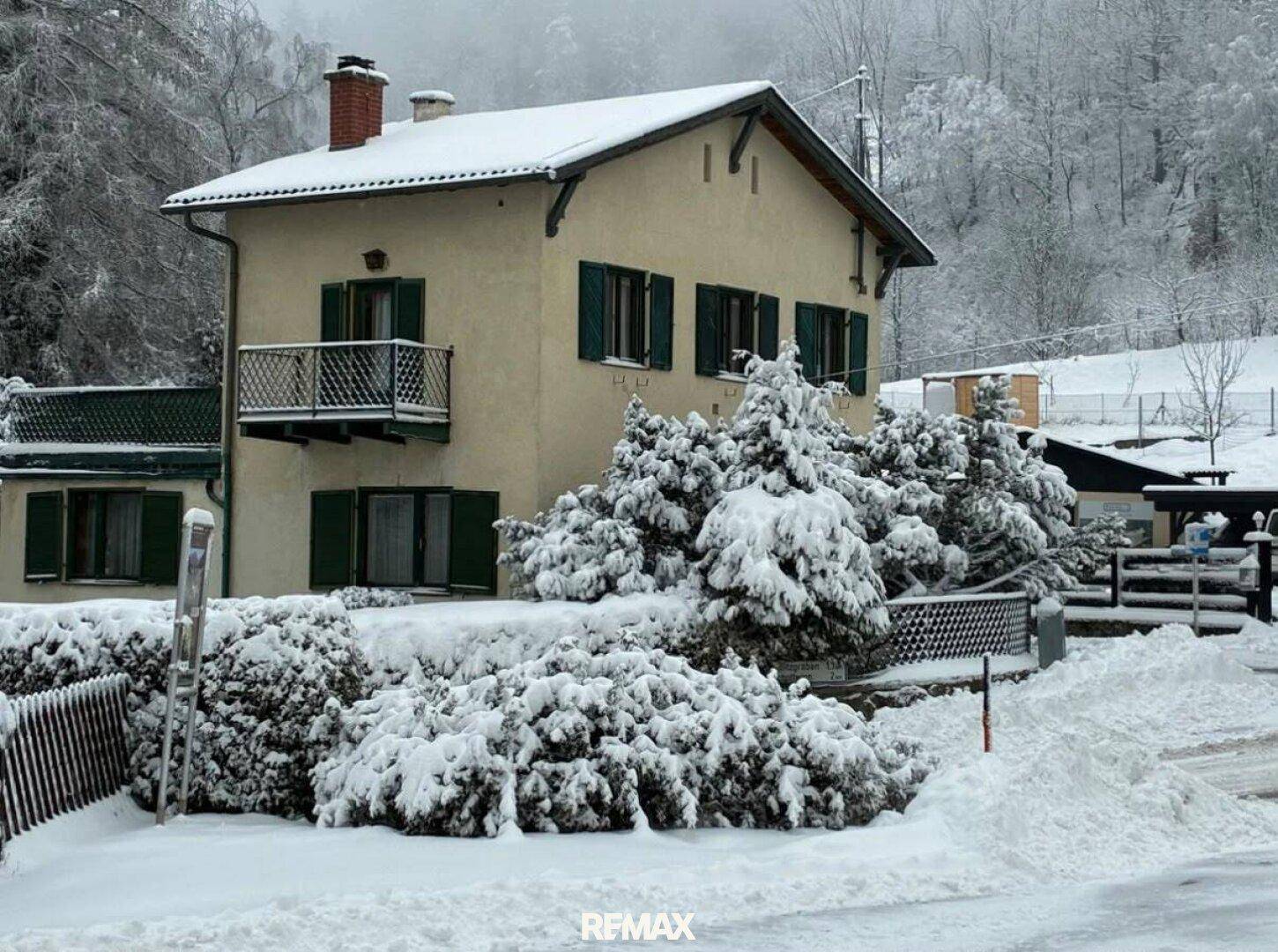 Winterliche Ansicht eines Hauses mit schneebedecktem Dach, grünen Fensterläden und kleinem Balkon.