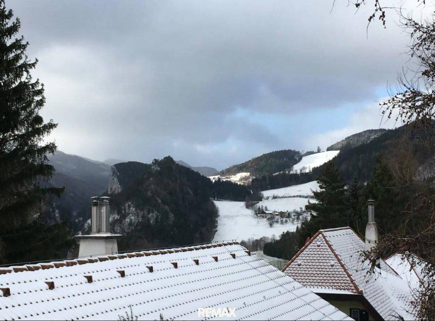 Weitblick über schneebedeckte Dächer und ein winterliches Tal mit bewaldeten Hügeln unter bewölktem Himmel.