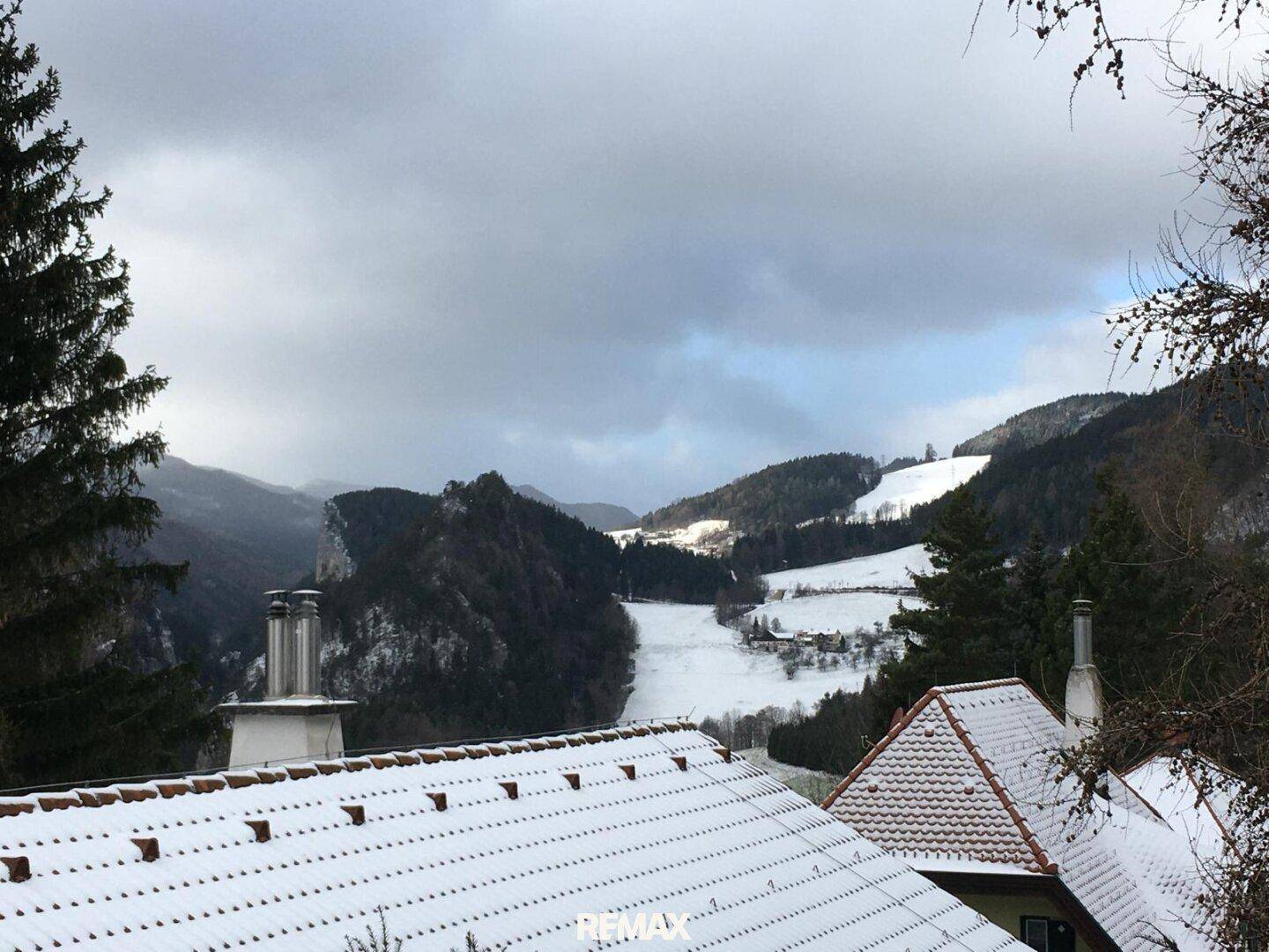Weitblick über schneebedeckte Dächer und ein winterliches Tal mit bewaldeten Hügeln unter bewölktem Himmel.