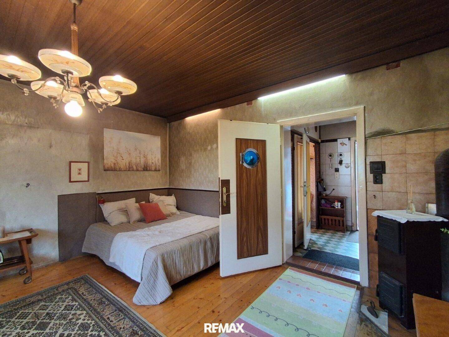 Schlafzimmer mit Holzboden, Bett und rustikaler Holzdeckenverkleidung, mit Zugang zu einem weiteren Raum.