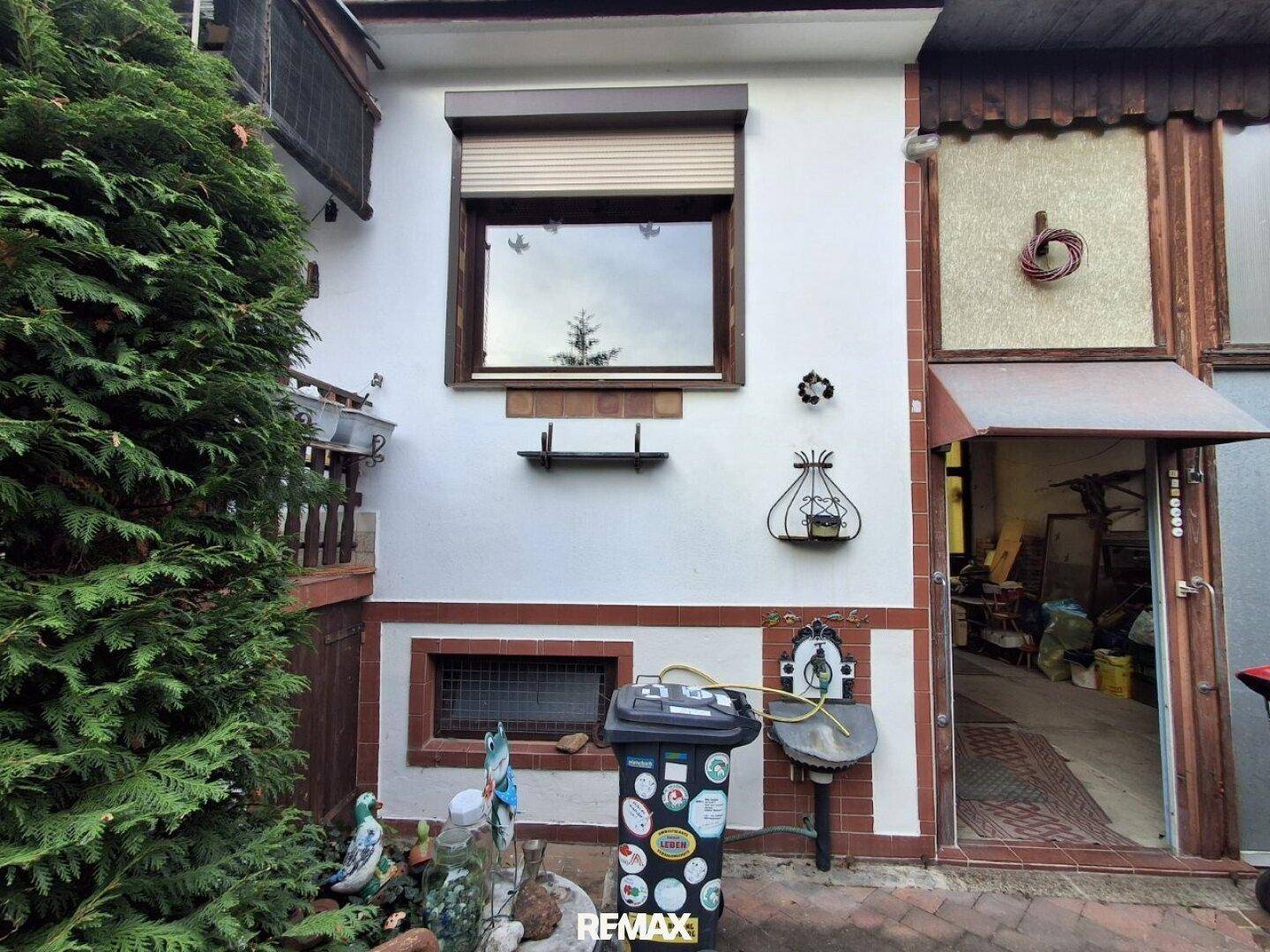 Außenansicht eines Gebäudes mit Fenster, Rollladen und einem Eingangsbereich, umgeben von Grün.
