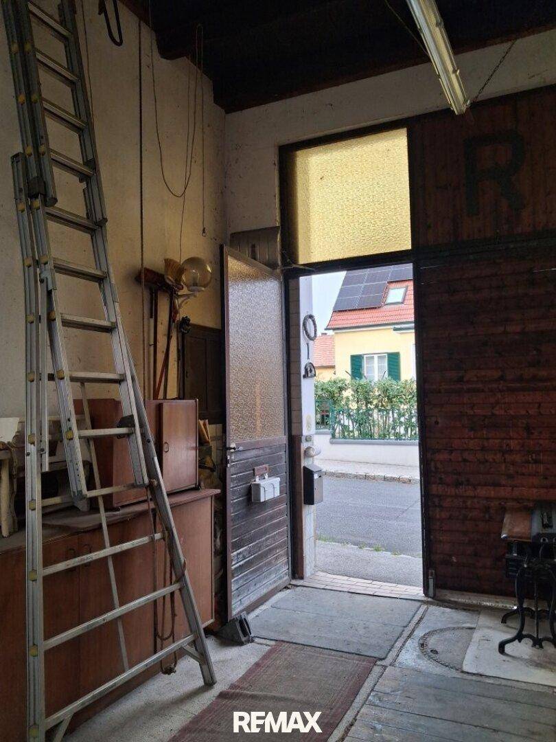 Innenraum mit Leiter und offenem Holztor, das den Blick auf ein Haus mit Solaranlage freigibt.