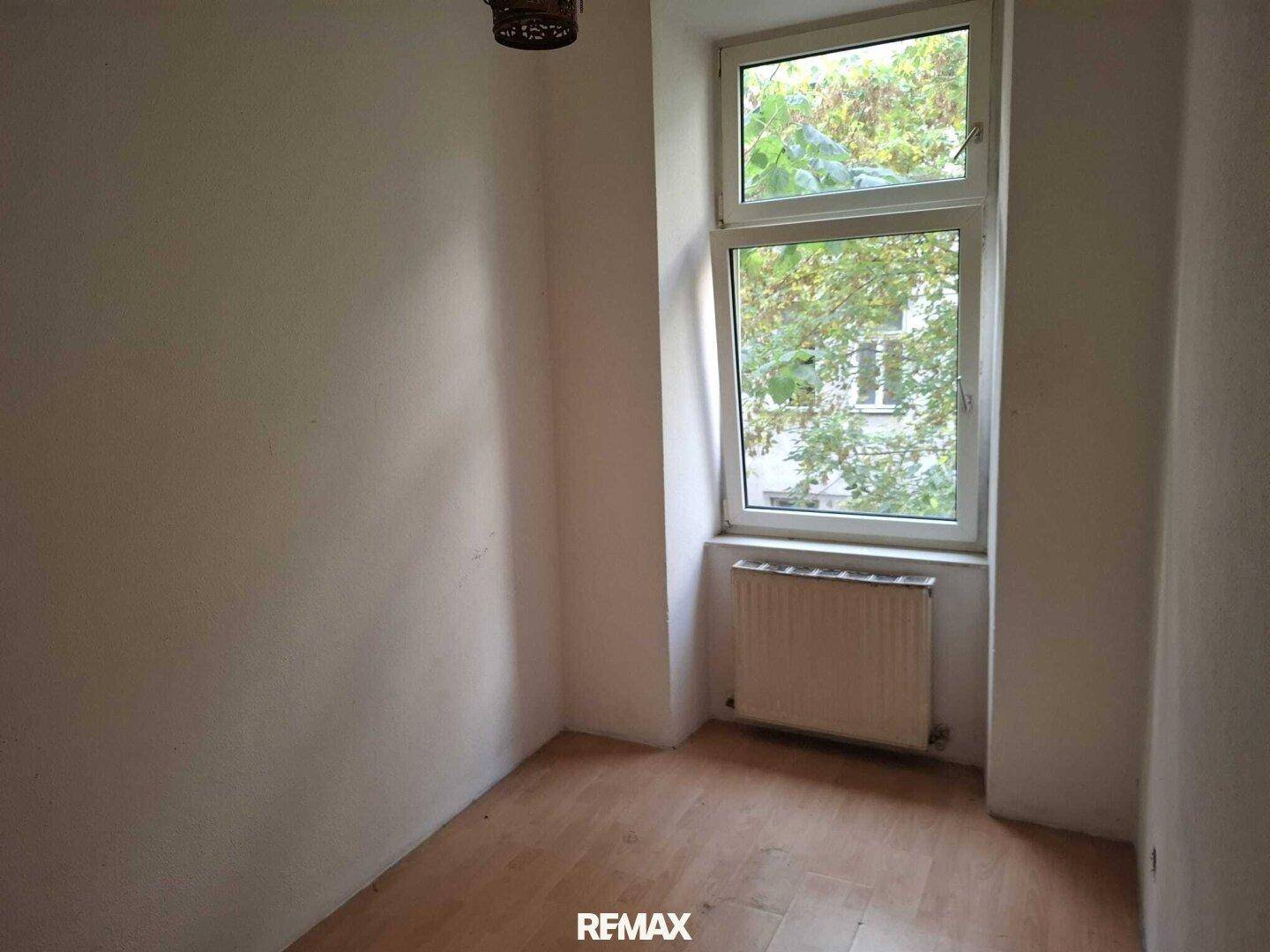 Kleines, leeres Zimmer mit Holzboden, einem Fenster und Heizkörper, in originalem Zustand.