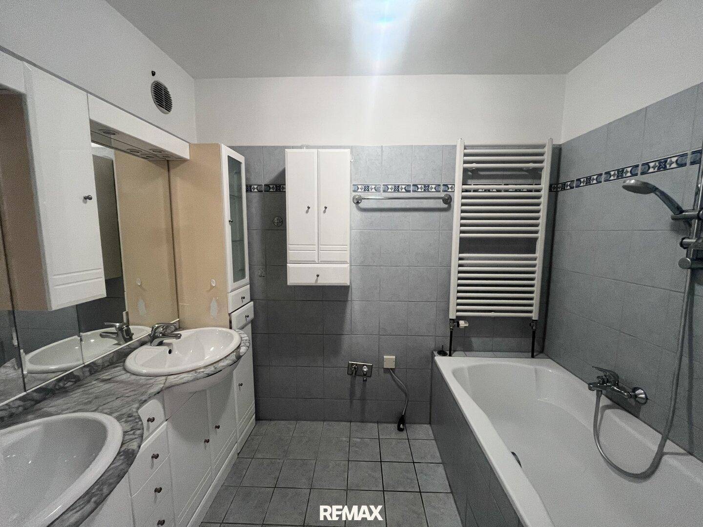 Badezimmer mit Badewanne, Doppelwaschbecken und gefliesten Wänden in Grau und Blau.