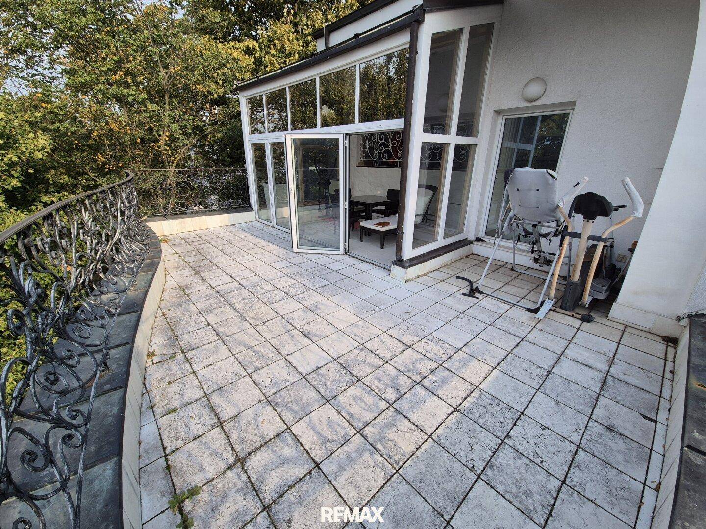 Geräumige geflieste Terrasse mit schmiedeeisernem Geländer und Zugang zu einem Wintergarten.