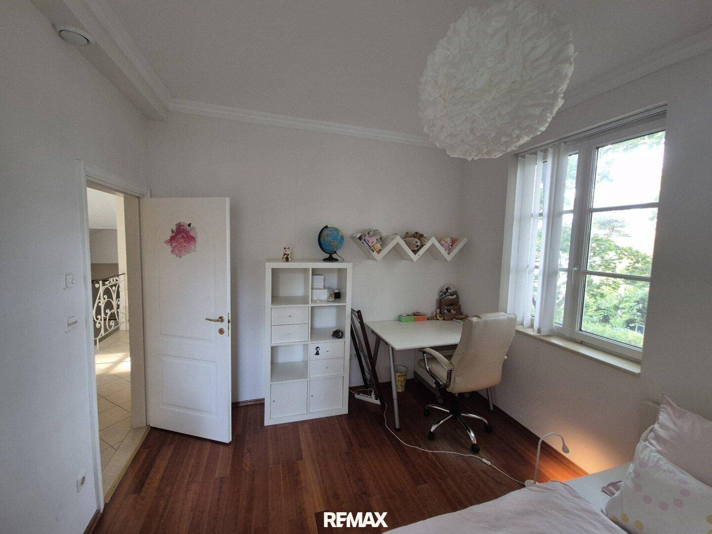Helles Schlafzimmer mit Holzfußboden, Schreibtisch und großem Fenster mit Grünblick.