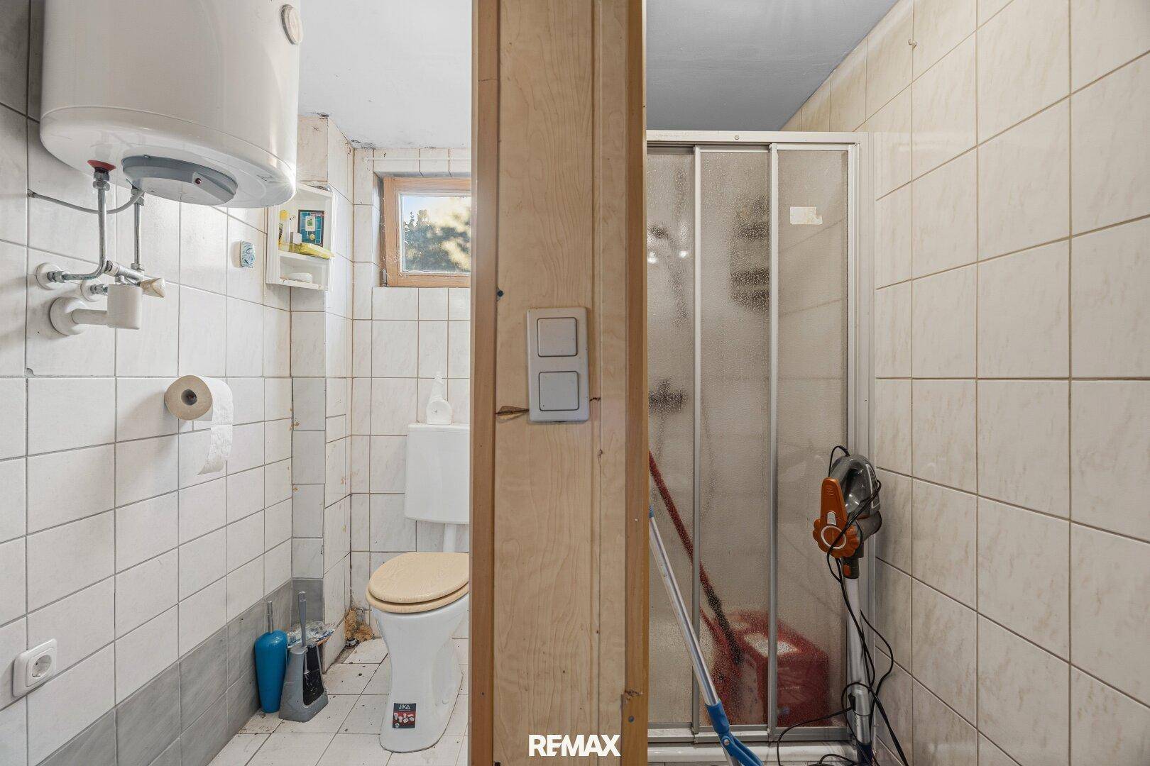 Einfaches Badezimmer mit weißen Fliesen, Toilette, Dusche und Warmwasserboiler.