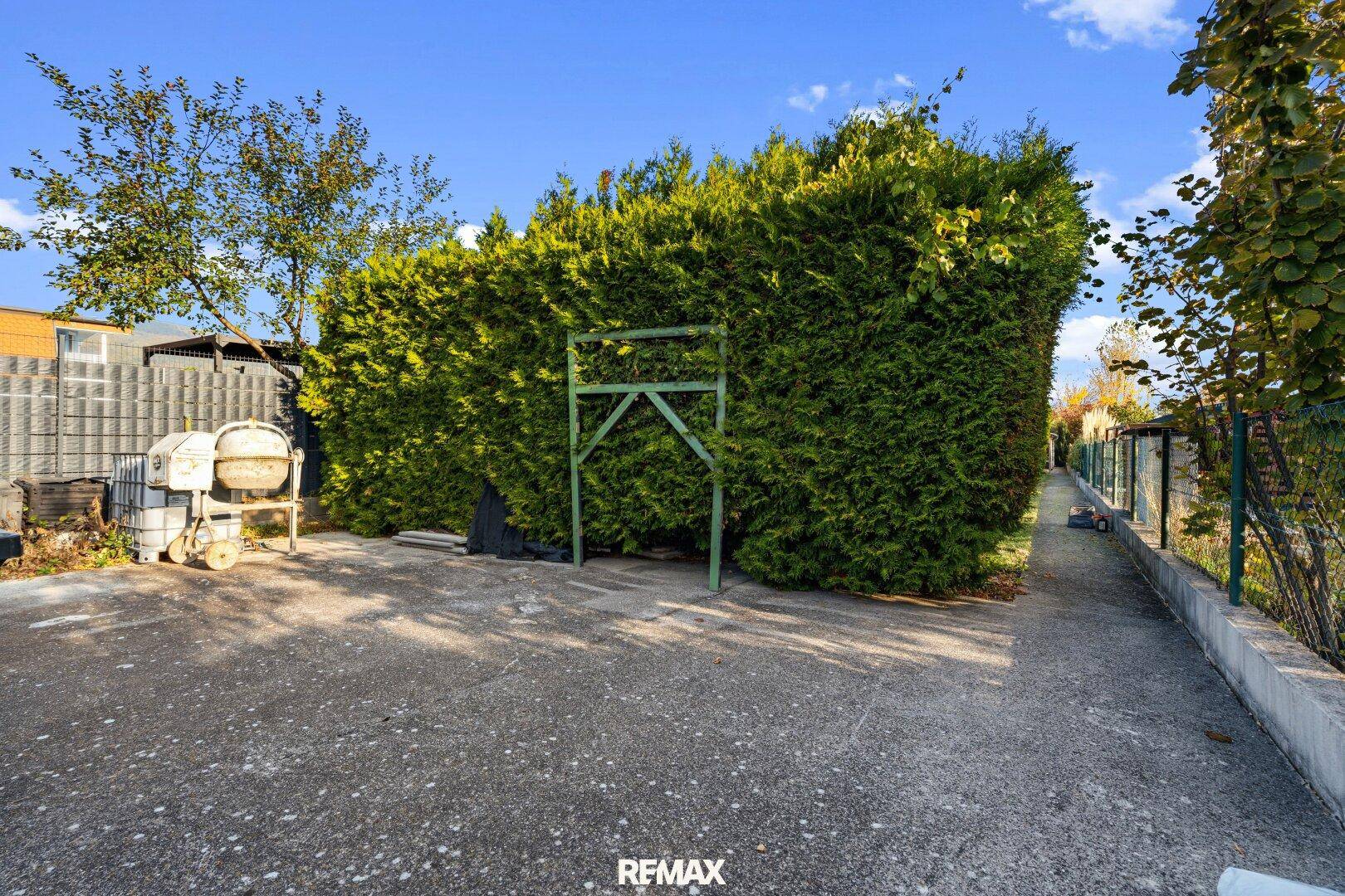 Gepflasterter Parkplatz mit hoher Hecke und einem schmalen Weg entlang des Grundstücks.