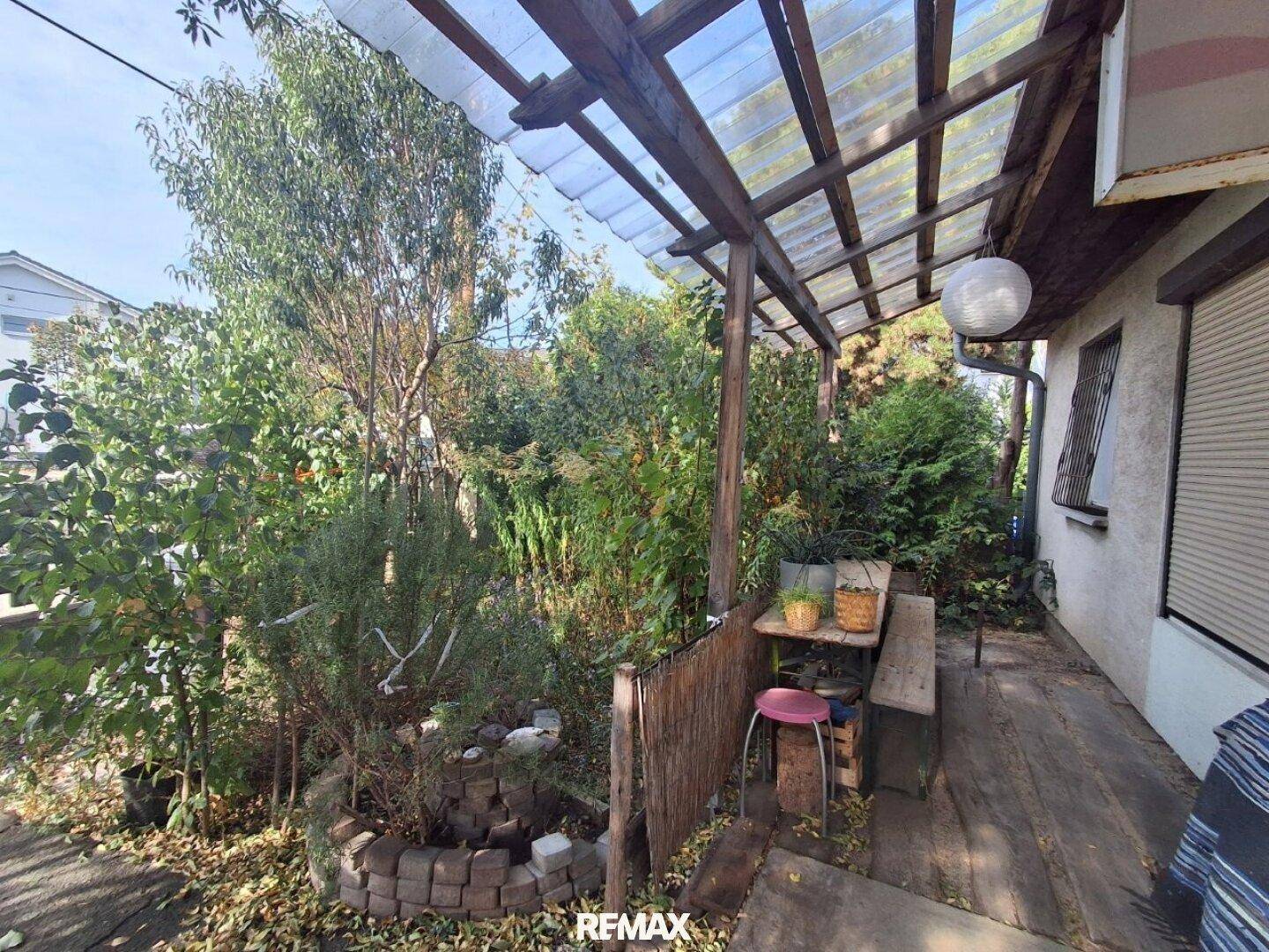 Überdachte Holzterrasse mit Sitzgelegenheiten, umgeben von üppigem Grün und Pflanzen.