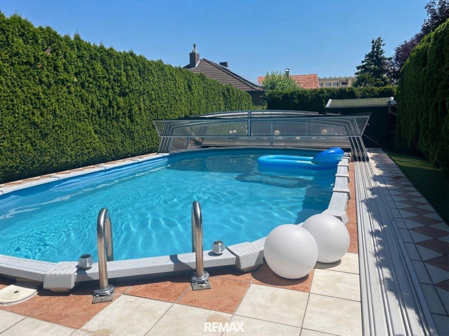 Außenpool mit Schiebeabdeckung und gepflegter Hecke im sonnigen Gartenbereich.