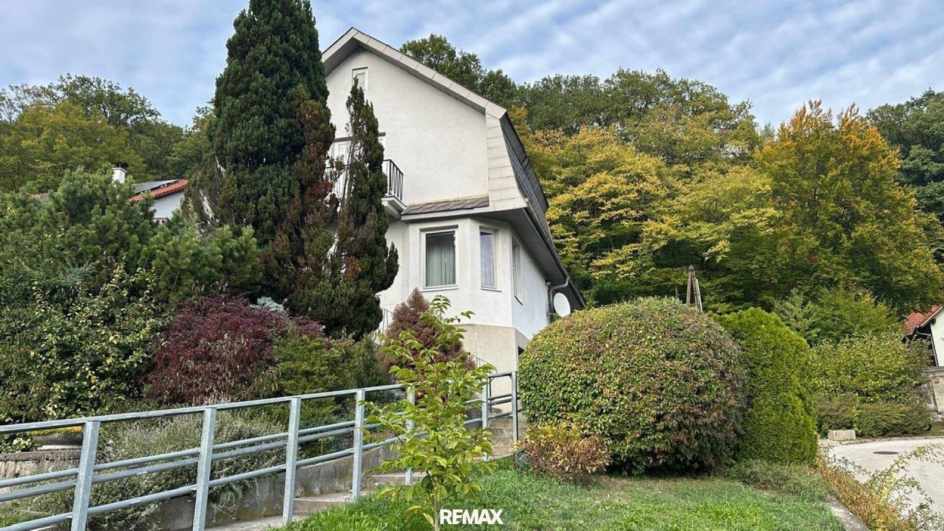 Freistehendes Haus mit Mansarddach, umgeben von Bäumen und Sträuchern auf einem Hanggrundstück.