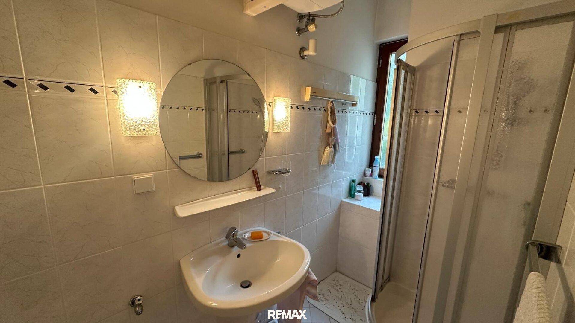Kleines Badezimmer mit Duschkabine, Waschbecken, Spiegel und hellen Fliesen an den Wänden.