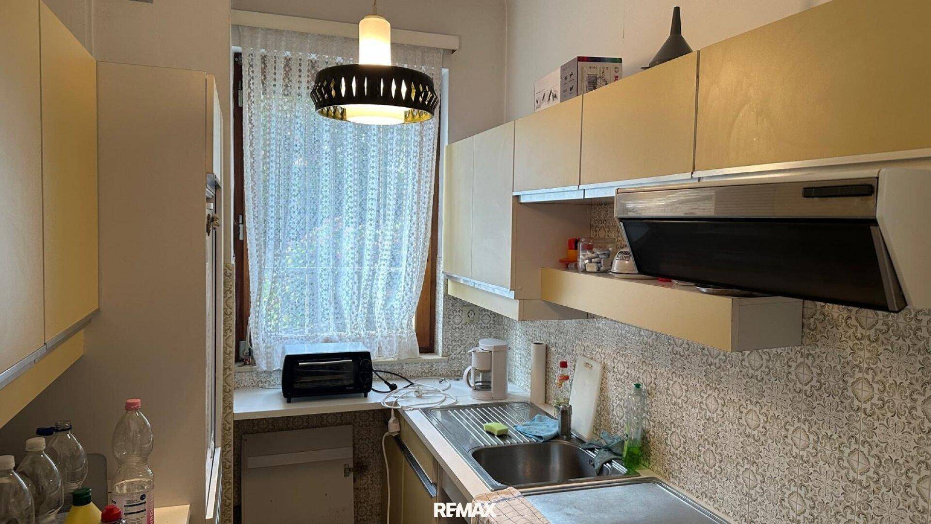 Helle Küche im Vintage-Stil mit beigen Schränken, Spüle und Fenster mit Vorhängen.