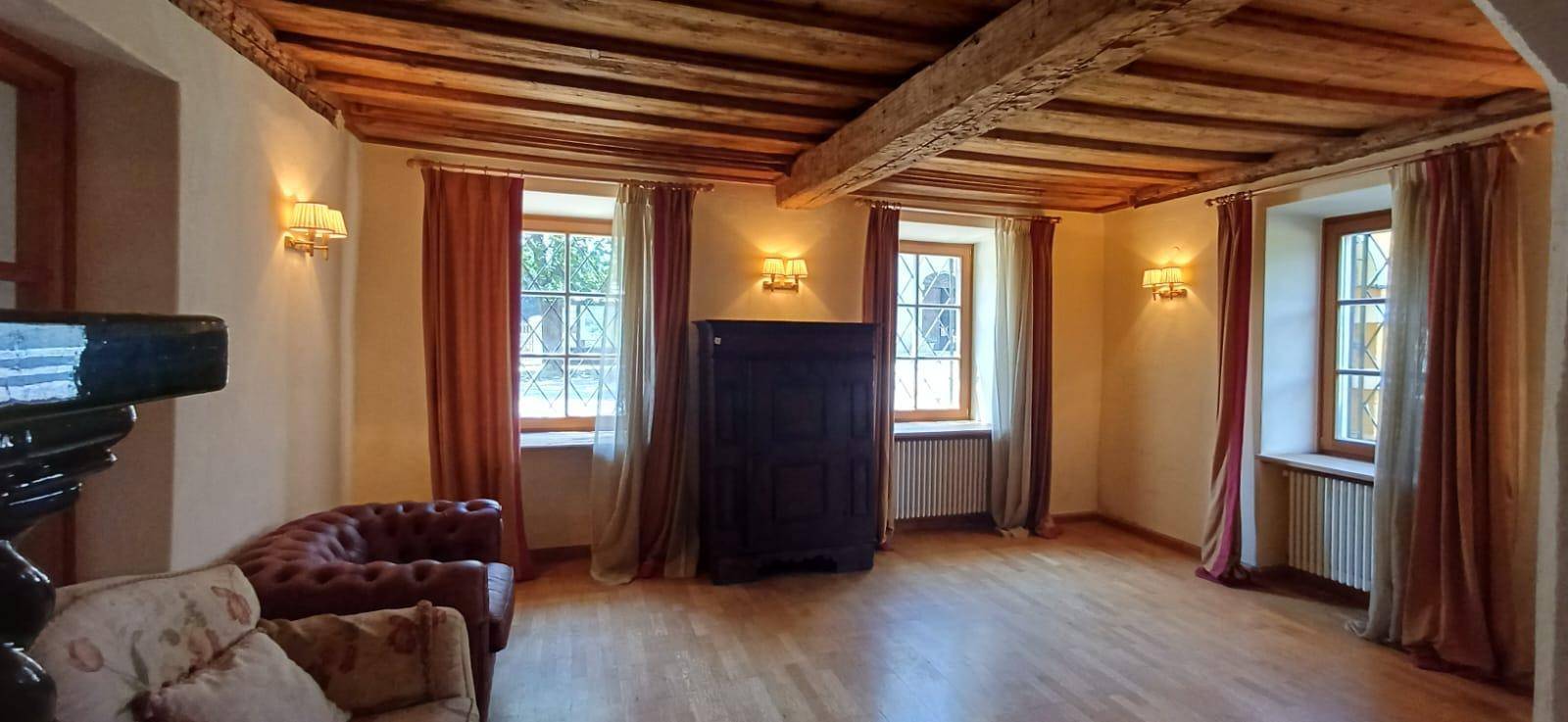 Heller Raum mit sichtbaren Holzbalken an der Decke, Holzboden und großen Fenstern mit Vorhängen.