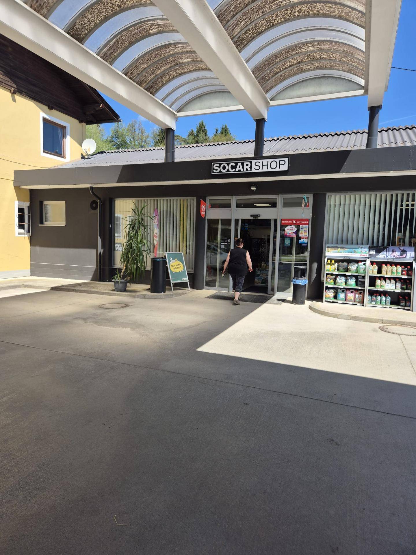 Eingangsbereich eines SOCAR Shops mit einer Person, die das Geschäft betritt, unter einem gewölbten Dach.