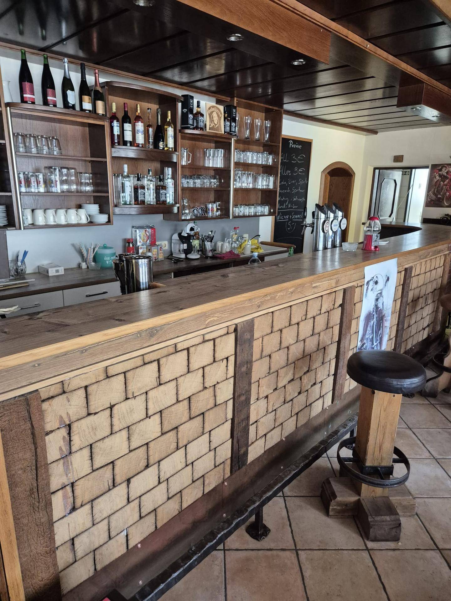 Rustikale Bar mit Holztheke und gefliestem Boden, dahinter Regale voller Flaschen und Gläser.