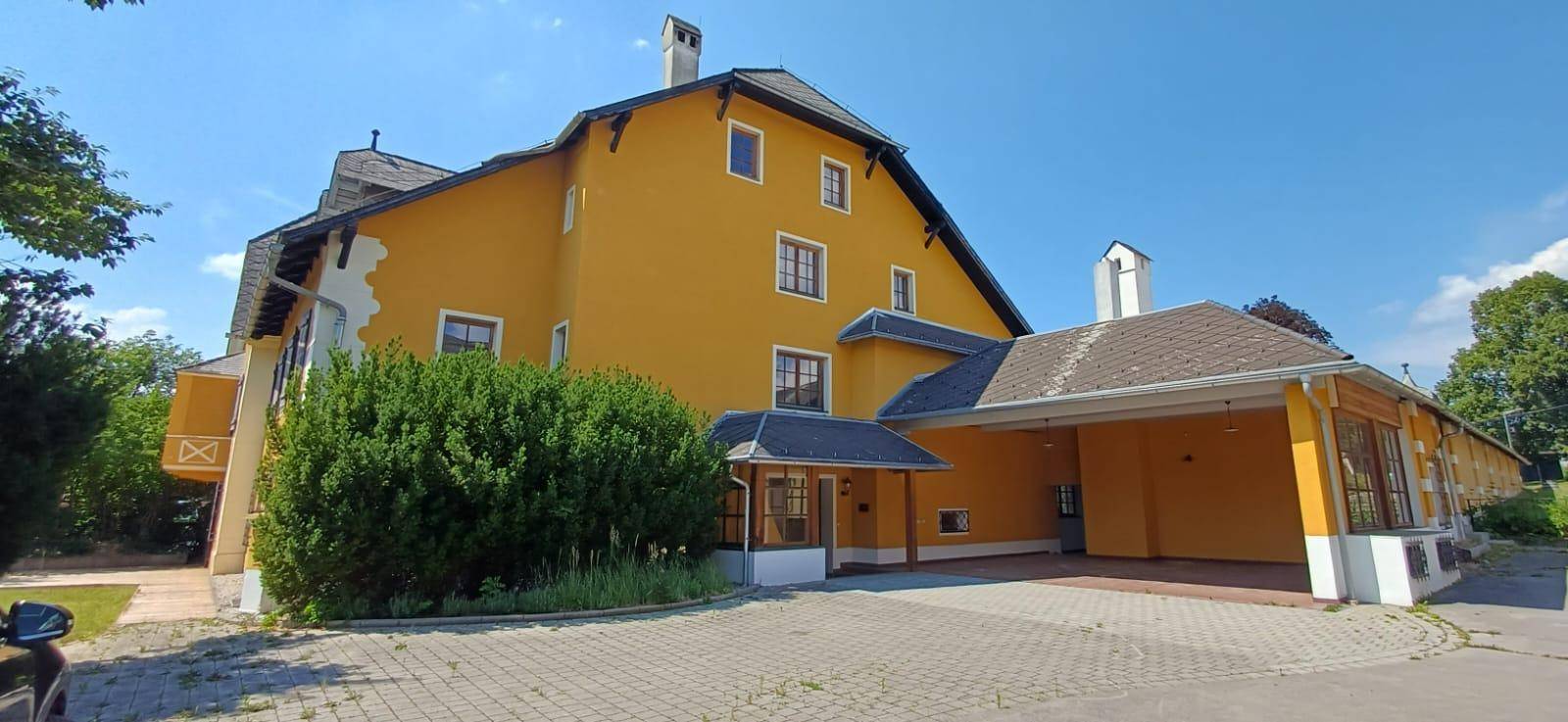 Seitenansicht des historischen Gebäudes mit gelber Fassade, Carport und gepflasterter Auffahrt unter blauem Himmel.