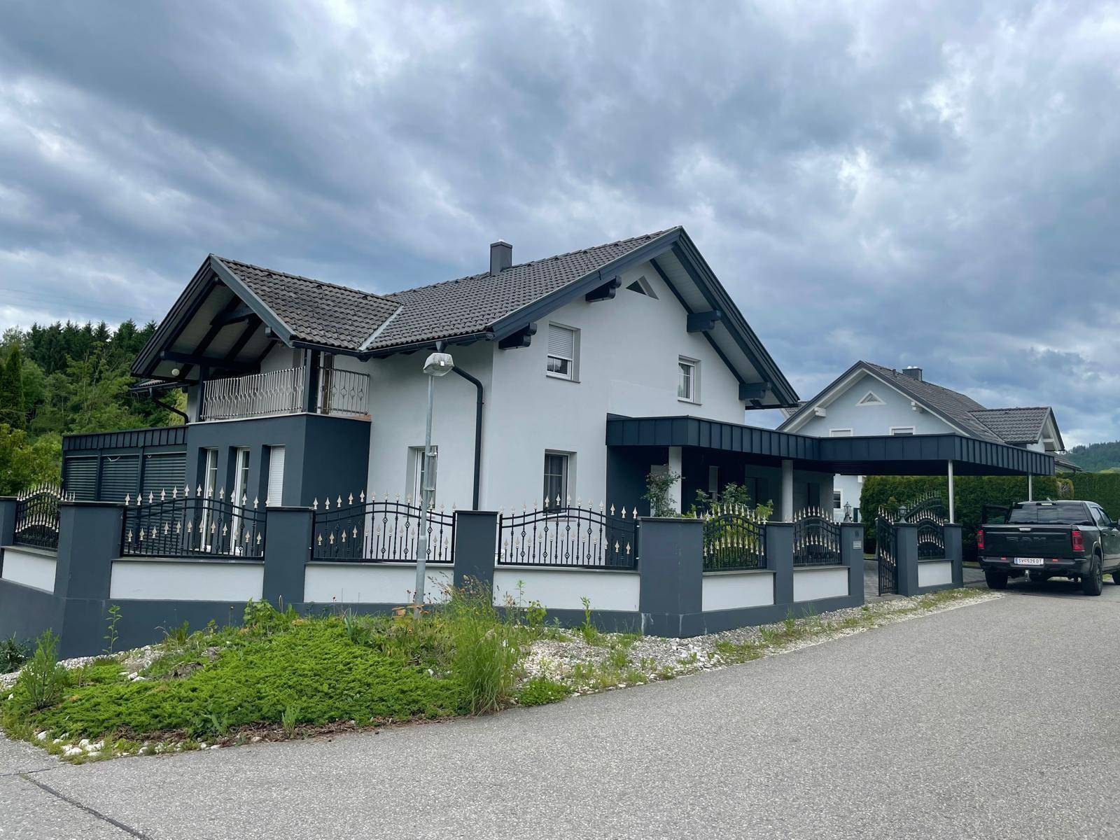 Seitenansicht des Hauses mit Zaun, Carport und gepflasterter Einfahrt unter bewölktem Himmel.