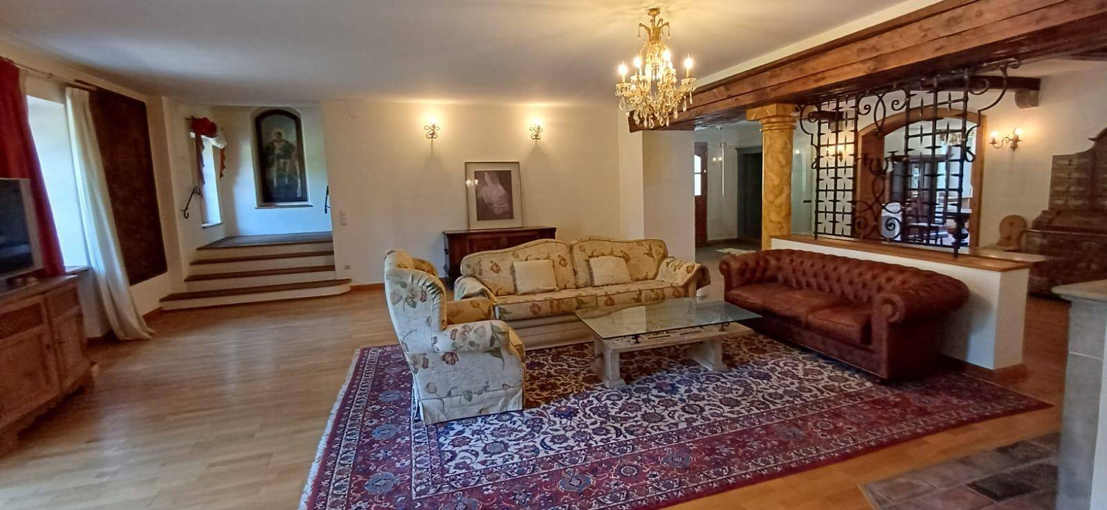 Weitläufiges Wohnzimmer mit eleganten Sofas, Kronleuchter und Blick auf eine Treppe in einem historischen Ambiente.