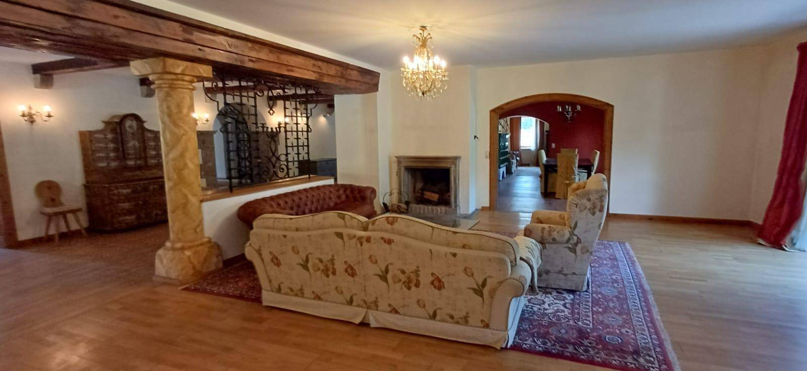 Großzügiges Wohnzimmer mit Kamin, Holzbalken und traditionellen Sofas, offen zu einem weiteren Bereich.