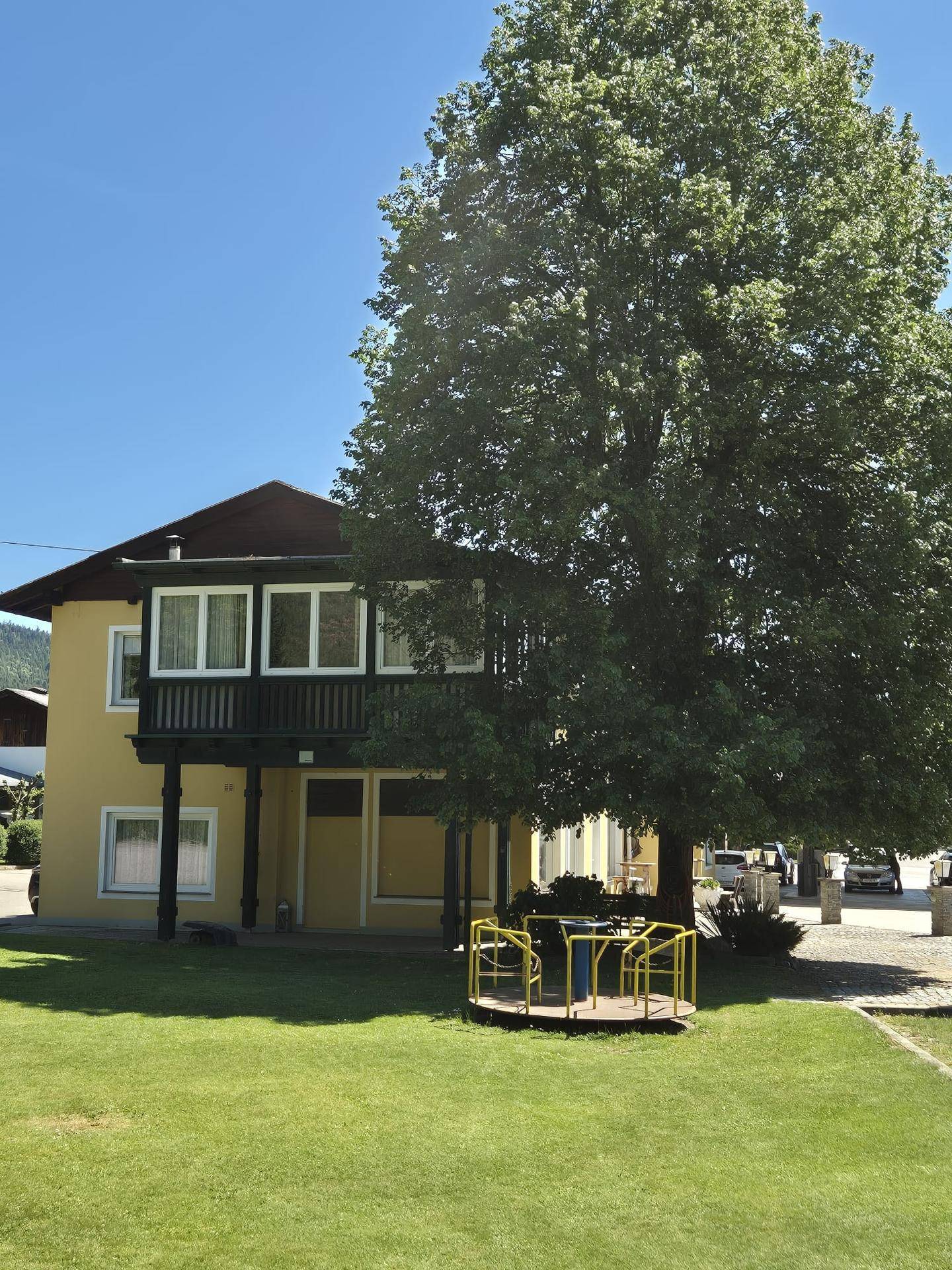 Gelbes Gebäude mit großem Baum und einem Karussell auf einer grünen Rasenfläche im Garten.