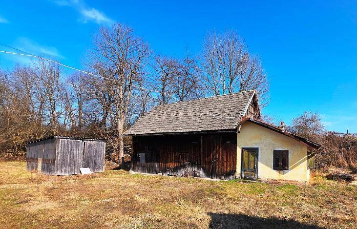 Ein älteres Gebäude mit Holzanbau und Schuppen, umgeben von trockener Wiese und Bäumen.