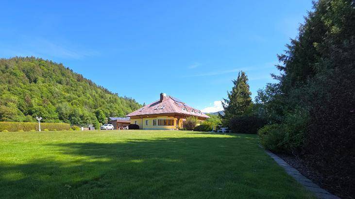 Gelbes Haus mit großem Garten und bewaldeten Hügeln im Hintergrund unter strahlend blauem Himmel.