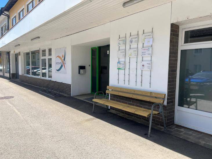 Eingangsbereich eines Gebäudes mit einer Holzbank, Informationsständern und großen Fenstern.