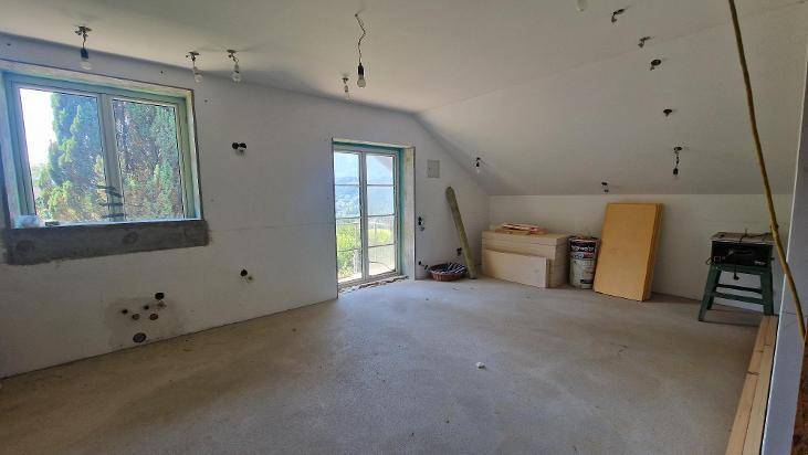 Unfertiger Dachbodenraum mit schräger Decke, Fenster und Zugang zu einem möglichen Balkon.