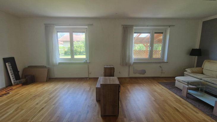 Heller Wohnbereich mit Holzboden, zwei Fenstern und einem beigen Sofa, teilweise renovierungsbedürftig.