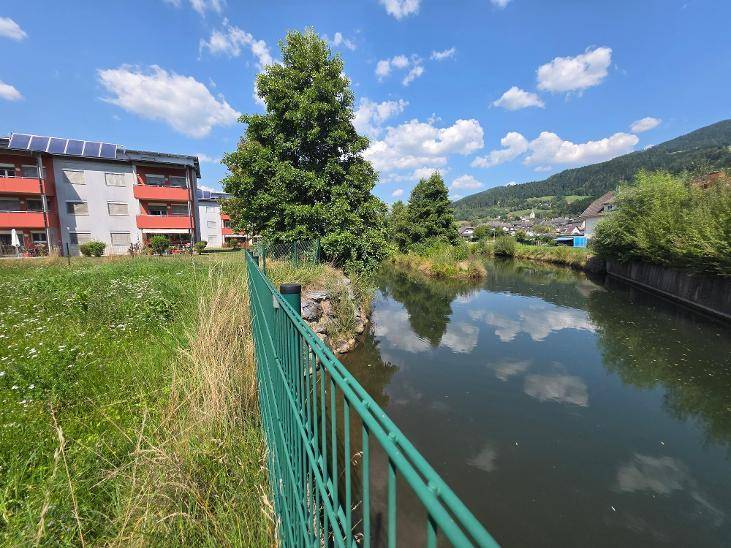 Flussufer mit grünem Zaun, angrenzendem Wohngebäude mit Solaranlagen und Bergblick.